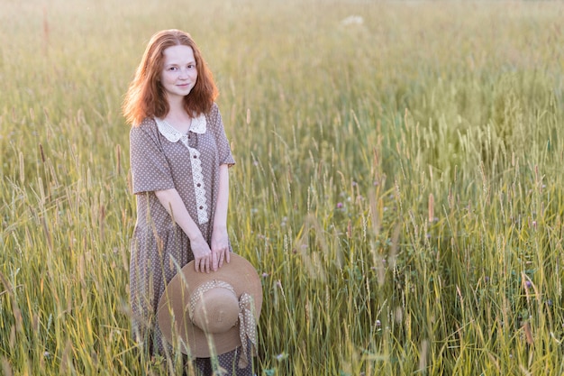 Redhead girl posing on a summer meadow at sunset in an elegant dress