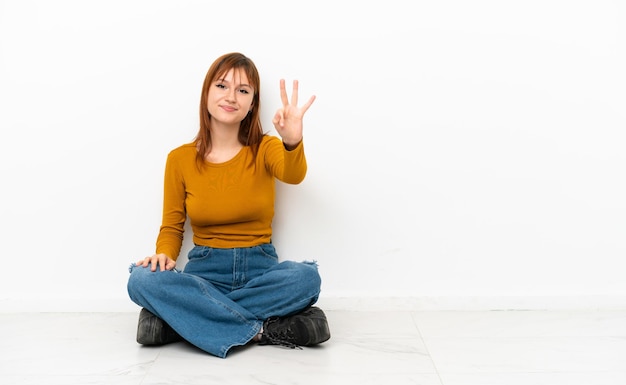 Redhead girl sitting on the floor isolated on white background happy and counting three with fingers