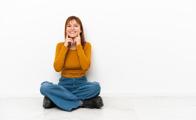 Foto ragazza rossa seduta sul pavimento isolato su sfondo bianco sorridente con un'espressione felice e piacevole