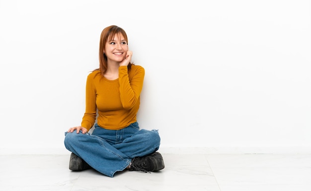 Photo redhead girl sitting on the floor isolated on white background thinking an idea while looking up