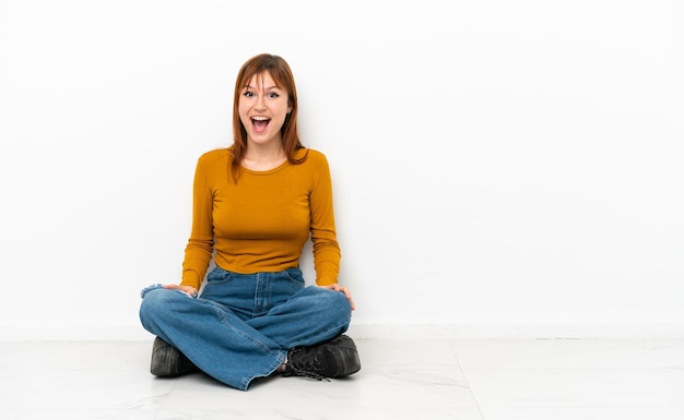 Redhead girl sitting on the floor isolated on white background with surprise facial expression