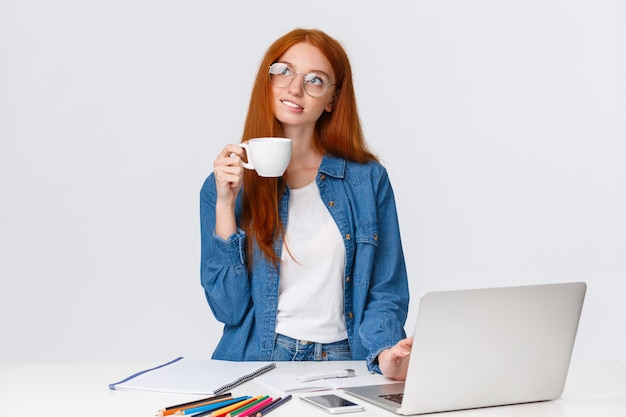 redhead girl standing with cup coffee near laptop and pencils