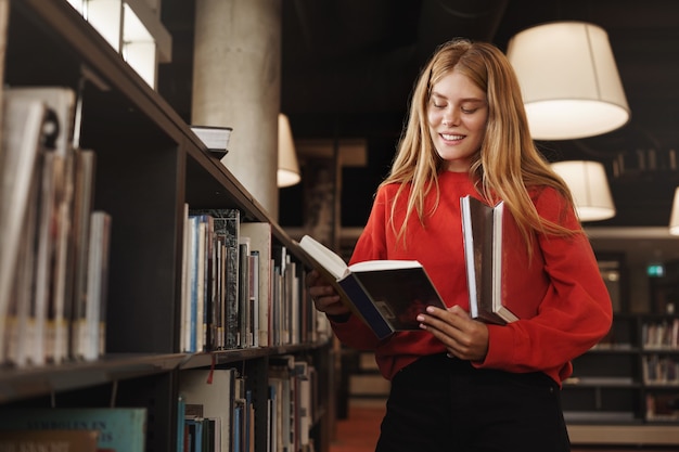 redhead girl, student standing in library near shelves, reading a book and smiling.