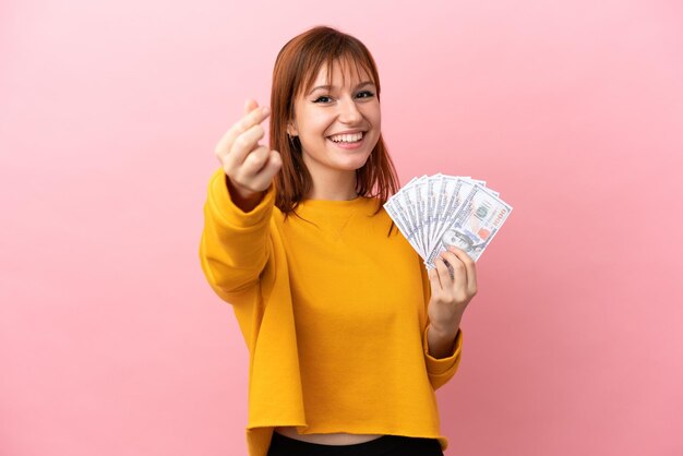 Redhead girl taking a lot of money isolated on pink background making money gesture