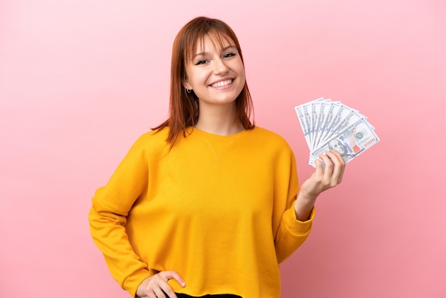 Redhead girl taking a lot of money isolated on pink background posing with arms at hip and smiling