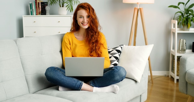 redhead girl using silver laptop while sitting on sofa in living room at home