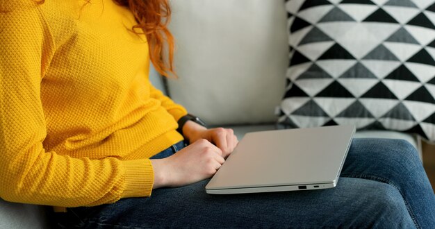 redhead girl using silver laptop while sitting on sofa in living room at home