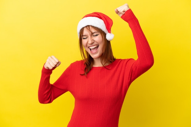 勝利を祝う黄色の背景に分離されたクリスマス帽子と赤毛の少女