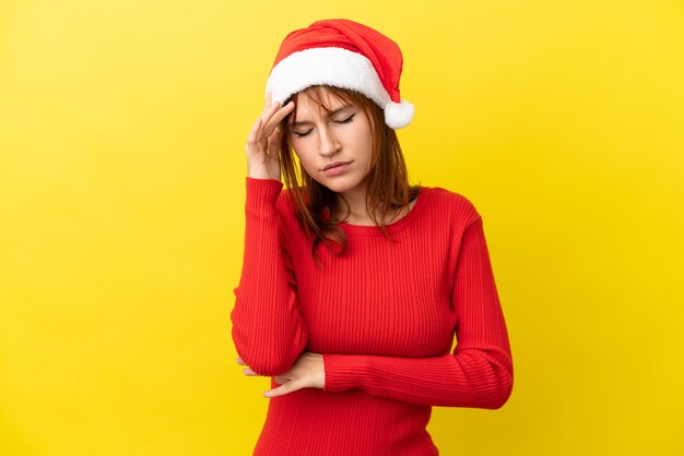 Redhead girl with christmas hat isolated on yellow background with headache