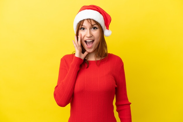 Redhead girl with christmas hat isolated on yellow background with surprise and shocked facial expression