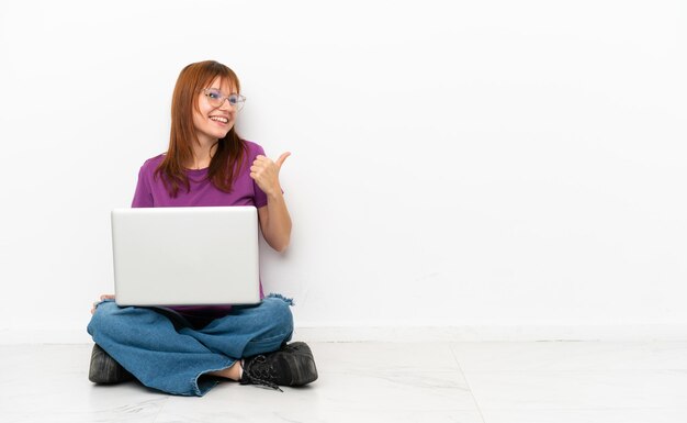 Redhead girl with a laptop sitting on the floor pointing to the side to present a product