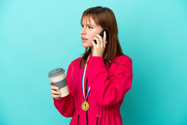 Redhead girl with medals isolated o blue background holding coffee to take away and a mobile