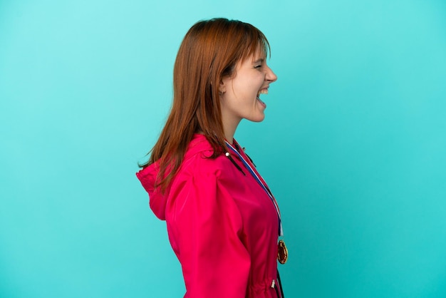 Redhead girl with medals isolated o blue background laughing in lateral position