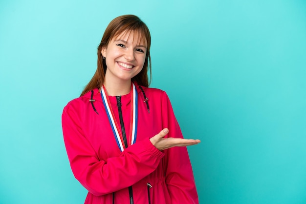 Redhead girl with medals isolated o blue background presenting an idea while looking smiling towards