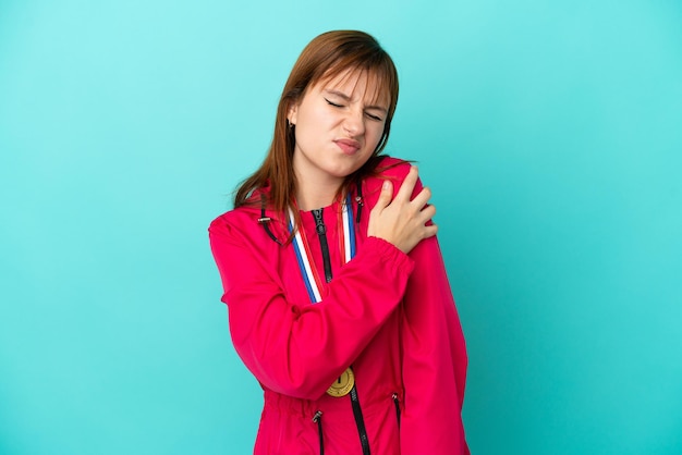 Redhead girl with medals isolated o blue background suffering from pain in shoulder for having made an effort