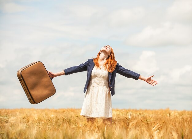 Redhead girl with suitcase at spring wheat field.