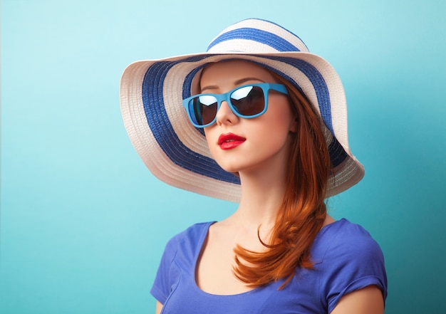 Redhead girl with sunglasses and hat on blue surface.