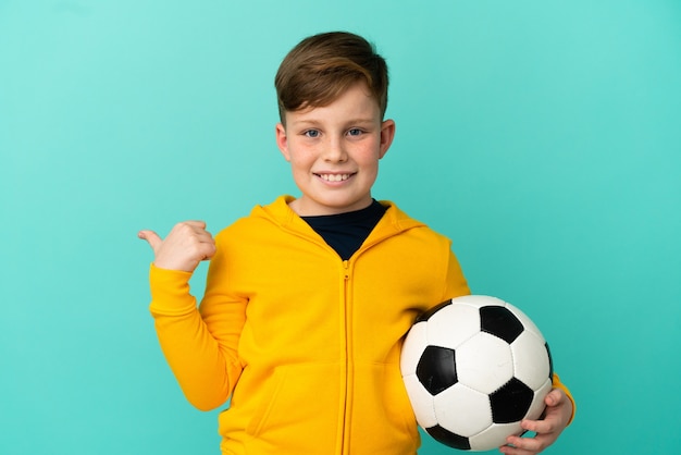 Redhead kid playing football isolated on blue background pointing to the side to present a product