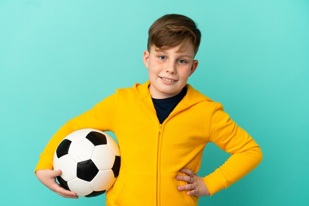 Ragazzo dai capelli rossi che gioca a calcio isolato su sfondo blu in posa con le braccia all'anca e sorridente