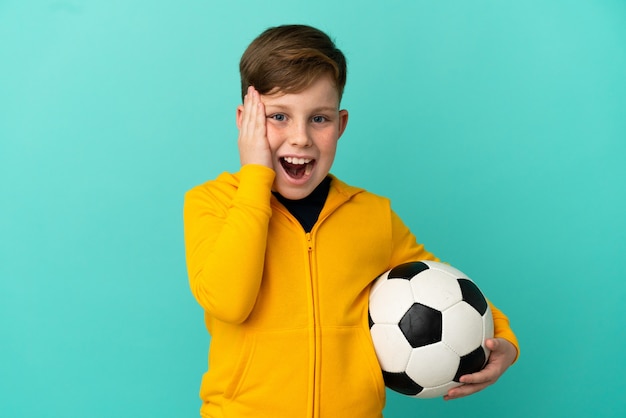 Redhead kid playing football isolated on blue background with surprise and shocked facial expression