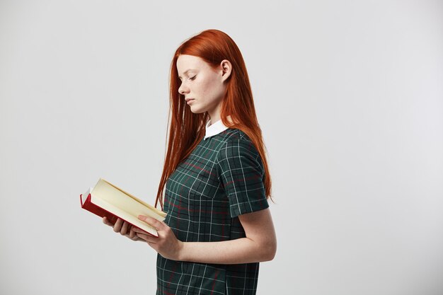 redhead long-haired girl in a green checkered dress reads a book on the white background 