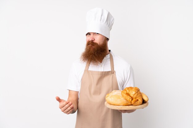 Redhead in uniforme da chef. panettiere maschio in possesso di un tavolo con diversi pani facendo dubbi gesto mentre si solleva le spalle