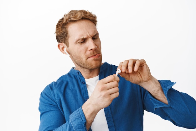 Redhead man listening music in wireless headphones, trying to clean earphone from dirt and frowning, standing over white background