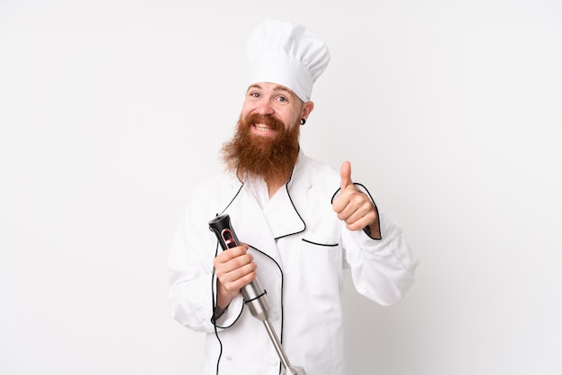 Redhead man using hand blender over isolated white wall giving a thumbs up gesture