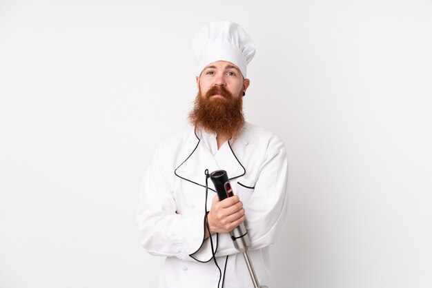 Redhead man using hand blender over isolated white wall keeping arms crossed