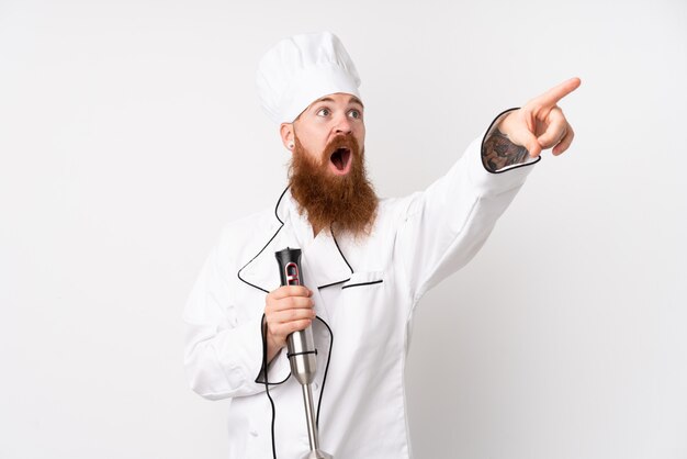 Redhead man using hand blender over isolated white wall pointing away