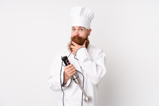 Redhead man using hand blender over white wall laughing