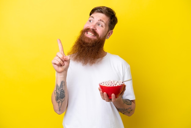 Рыжий мужчина с бородой ест миску хлопьев, изолированных на желтом фоне, намереваясь реализовать решение, поднимая палец вверх