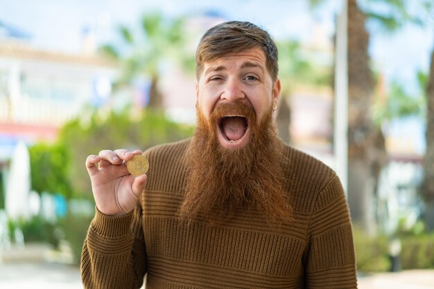 Photo redhead man with beard holding a bitcoin at outdoors with surprise and shocked facial expression