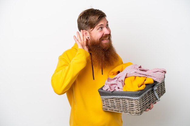 Redhead man with beard holding a clothes basket isolated on white background listening to something by putting hand on the ear