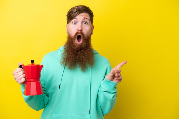 Рыжий мужчина с бородой, держащий кофейник на желтом фоне, удивлен и указывает в сторону