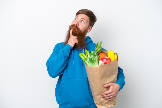 白い背景に隔離された食料品の買い物袋を持ち、見上げるひげを持つ赤毛の男