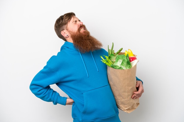 努力をしたために腰痛に苦しんでいる白い背景に分離された食料品の買い物袋を保持しているひげを持つ赤毛の男