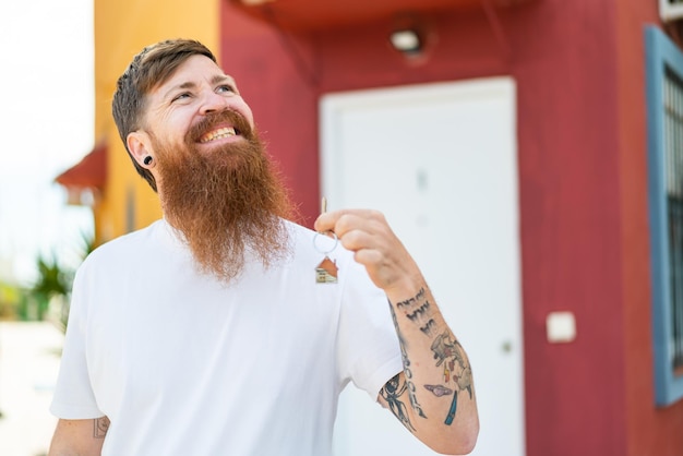 Redhead man with beard holding home keys at outdoors looking up while smiling