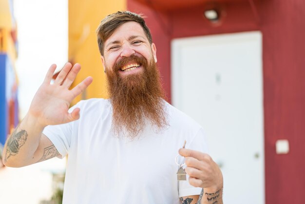 Photo redhead man with beard holding home keys at outdoors saluting with hand with happy expression