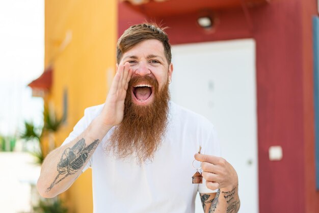 Photo redhead man with beard holding home keys at outdoors shouting with mouth wide open