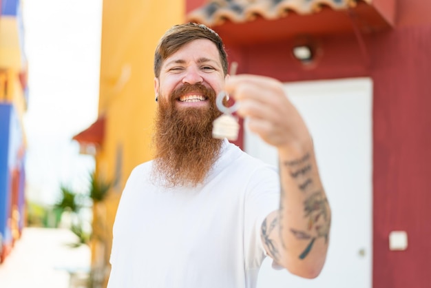 Redhead man with beard holding home keys at outdoors with happy expression