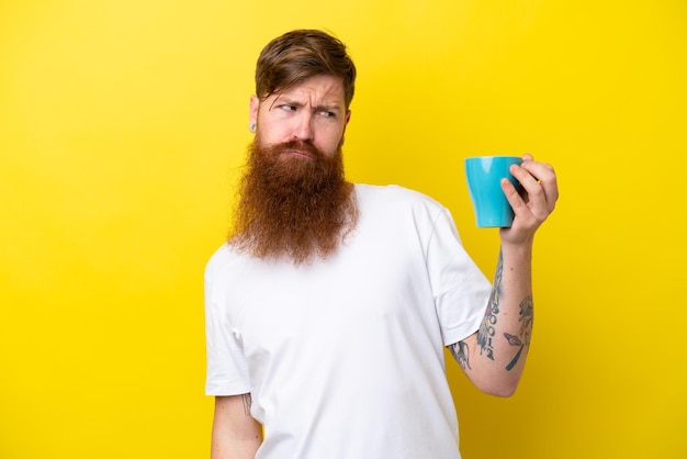 Redhead man with beard holding a mug isolated on yellow background with sad expression