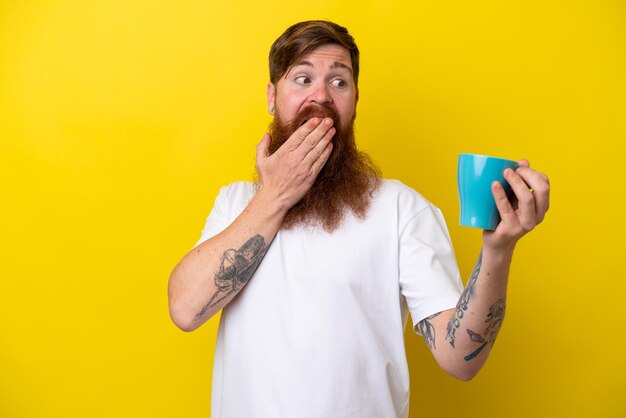 Redhead man with beard holding a mug isolated on yellow background with surprise and shocked facial expression