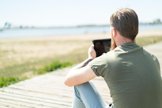 Redhead man with beard holding a tablet at outdoors in back position