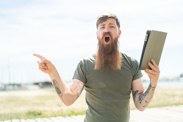 Redhead man with beard holding a tablet at outdoors surprised and pointing side