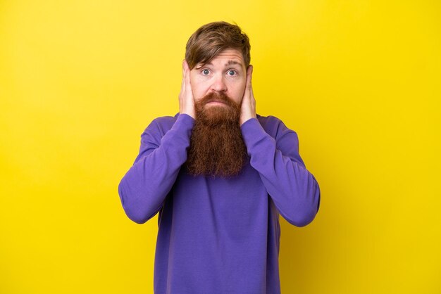 Фото Рыжий мужчина с бородой на желтом фоне расстроен и закрывает уши