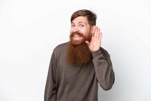 Redhead man with beard isolated on white background listening to something by putting hand on the ear