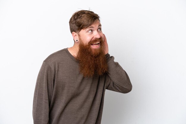 Redhead man with beard isolated on white background listening to something by putting hand on the ear