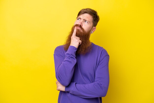 Redhead man with beard isolated on yellow background having doubts while looking up