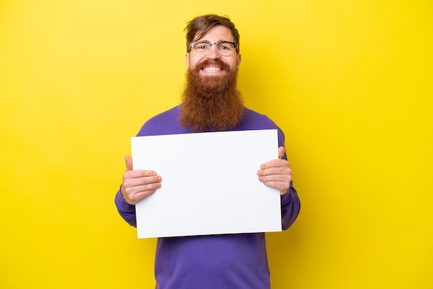 Redhead man with beard isolated on yellow background holding an empty placard with happy expression